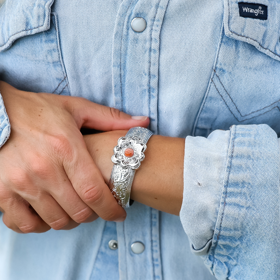 A person in a light denim Wrangler shirt is wearing The Eloise Cuff—a silver floral bracelet with an orange stone—holding their wrist with the opposite hand. It's a striking piece of statement jewelry that adds a bold Western flair.