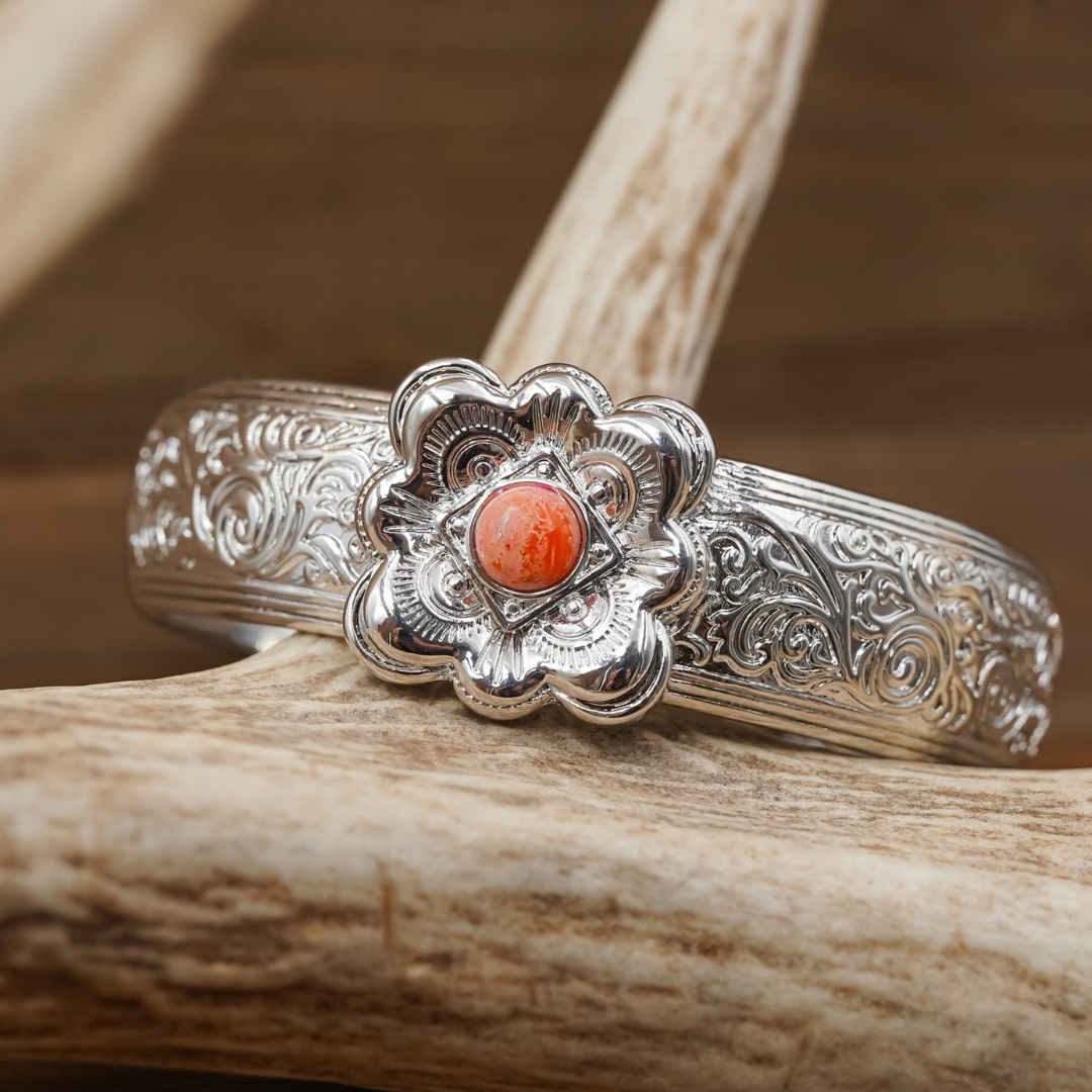The Eloise Cuff is a silver bracelet with ornate floral engravings that recall the classic Western cuff style, highlighted by a central flower-shaped setting featuring an orange-red gemstone, elegantly displayed on light brown antlers.