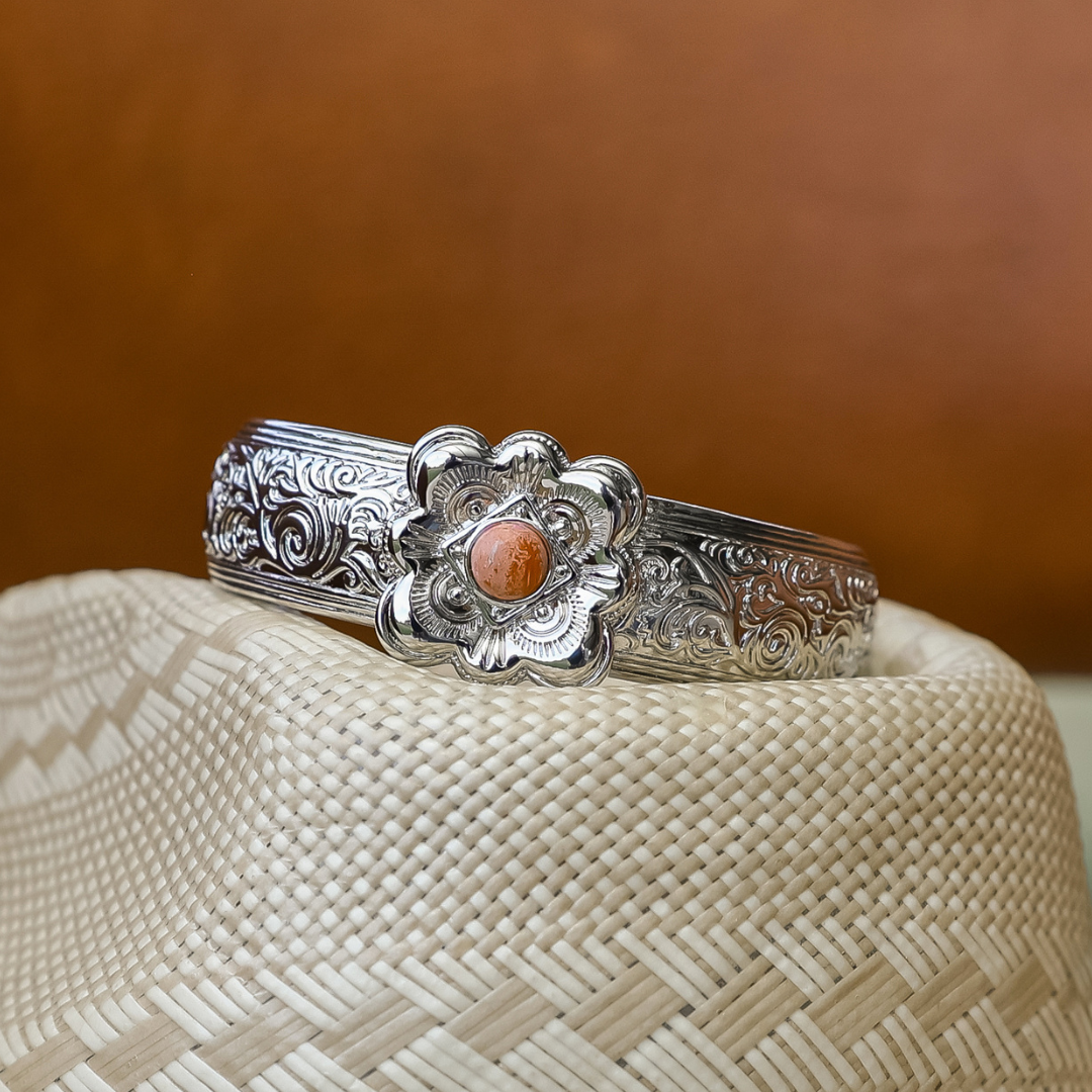 The Eloise Cuff, a silver bracelet featuring ornate floral carvings and a striking central orange stone, rests elegantly on a woven straw hat, adding the perfect statement jewelry accent against a warm brown background.