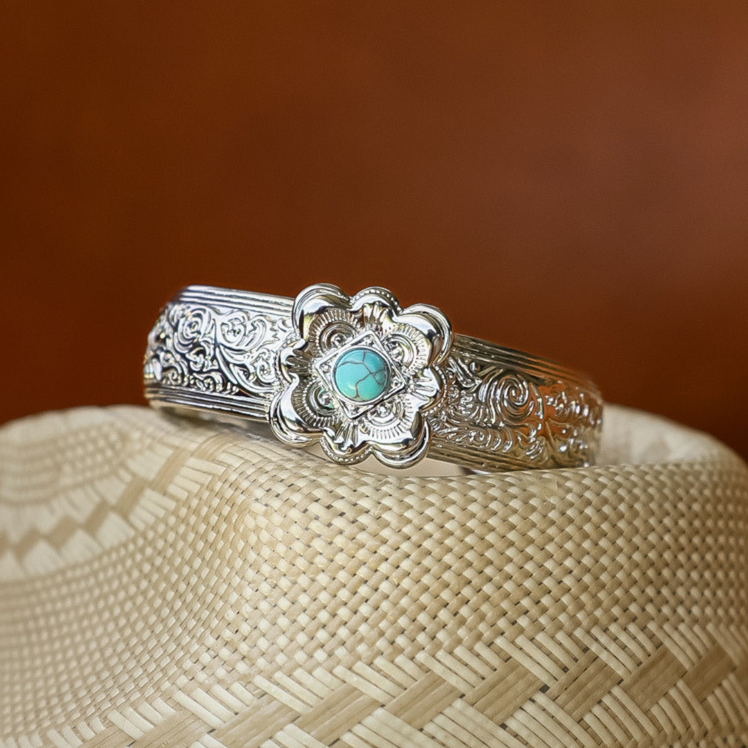 The Eloise Cuff—a silver bracelet adorned with ornate floral engravings and a turquoise stone centerpiece—rests atop a woven straw hat, embodying the essence of statement jewelry and evoking the timeless charm of a Western cuff.