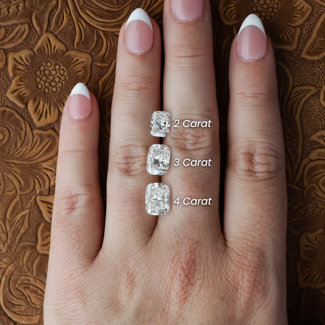 A hand with a French manicure showcases three dazzling cushion-cut diamonds—including the 2 ct. Elongated Cushion Cut Lab Grown Diamond—each labeled 2 carat, 3 carat, and 4 carat on the middle finger, set against a floral patterned brown surface.