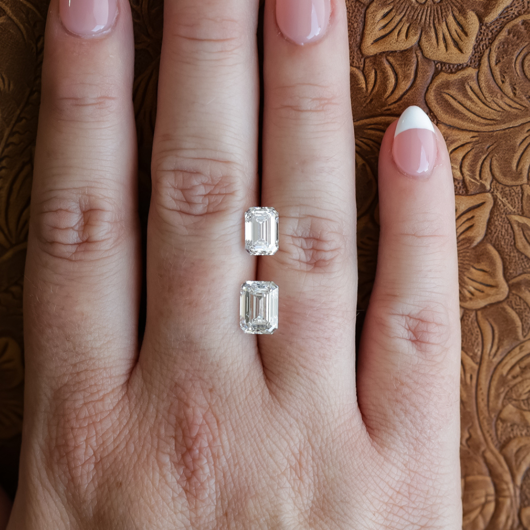 A hand with a French manicure wears a ring featuring two 2 ct. Emerald Cut Lab Grown Diamonds, set parallel on the middle finger, against a textured brown background. Each IGI certified diamond sparkles brilliantly, highlighting the elegant sophistication of this modern design.
