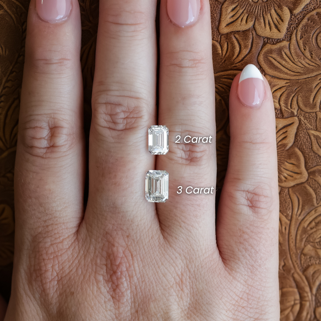 A hand displays two emerald-cut diamond rings: a 2 ct. Emerald Cut Lab Grown Diamond on the middle finger and a 3-carat emerald-cut lab-grown diamond on the ring finger, each labeled accordingly. The nails are manicured with light pink polish.