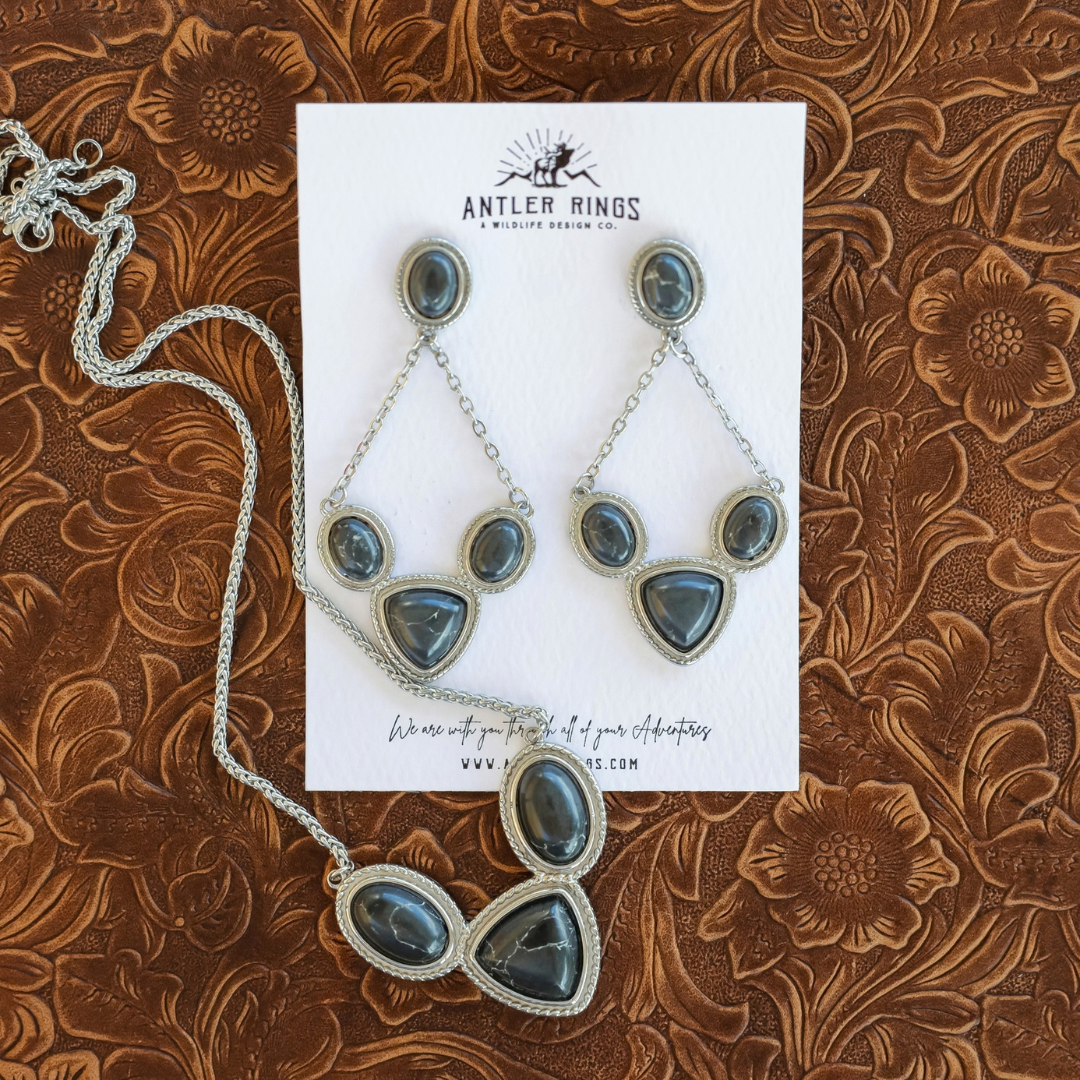 The Eve Drop Earrings, a pair of silver drop earrings with three gray stone cabochons each, displayed on a card next to a matching Western necklace.