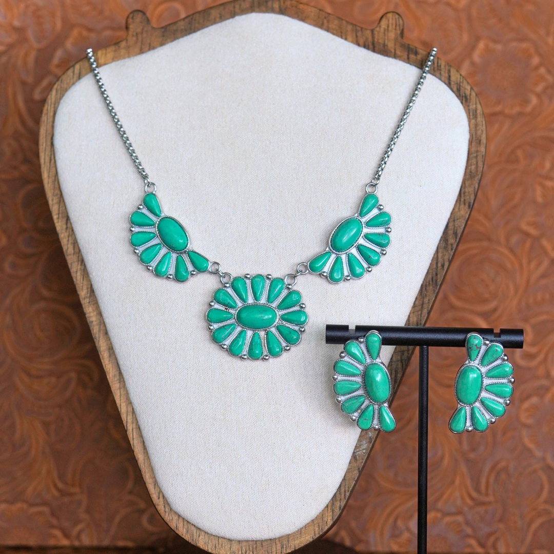 A silver necklace and The Fern Stud Earrings, featuring green oval stones in floral patterns, are displayed on a cream jewelry stand against a brown textured background.