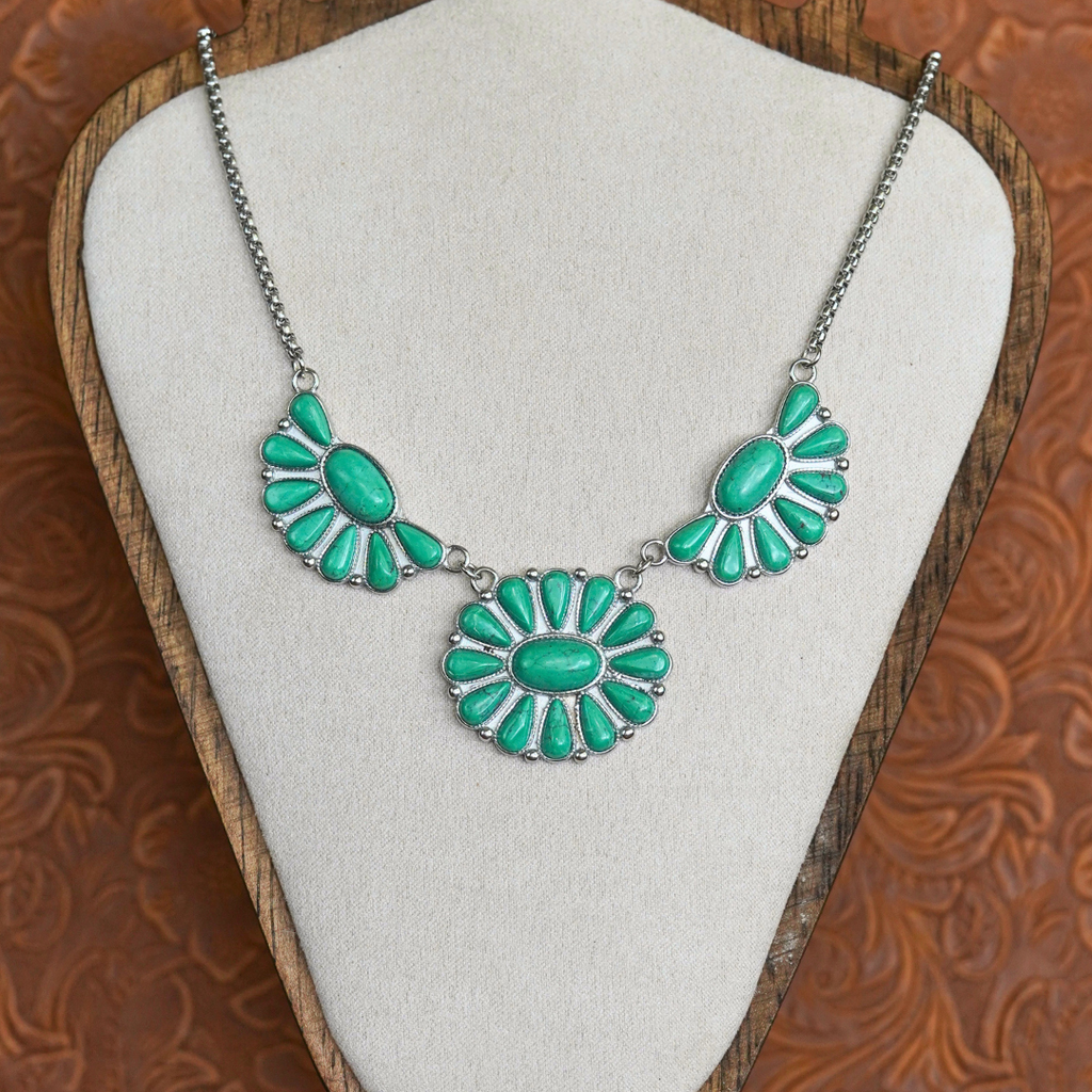 The Fern, a stunning Western necklace featuring oval turquoise stones set in three silver floral patterns, is showcased on a beige fabric bust with a wooden frame and an embossed brown backdrop—making it the perfect accessory for fans of classic cowgirl jewelry.