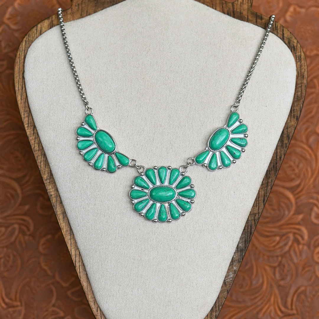 The Fern, a stunning Western necklace featuring oval turquoise stones set in three silver floral patterns, is showcased on a beige fabric bust with a wooden frame and an embossed brown backdrop—making it the perfect accessory for fans of classic cowgirl jewelry.