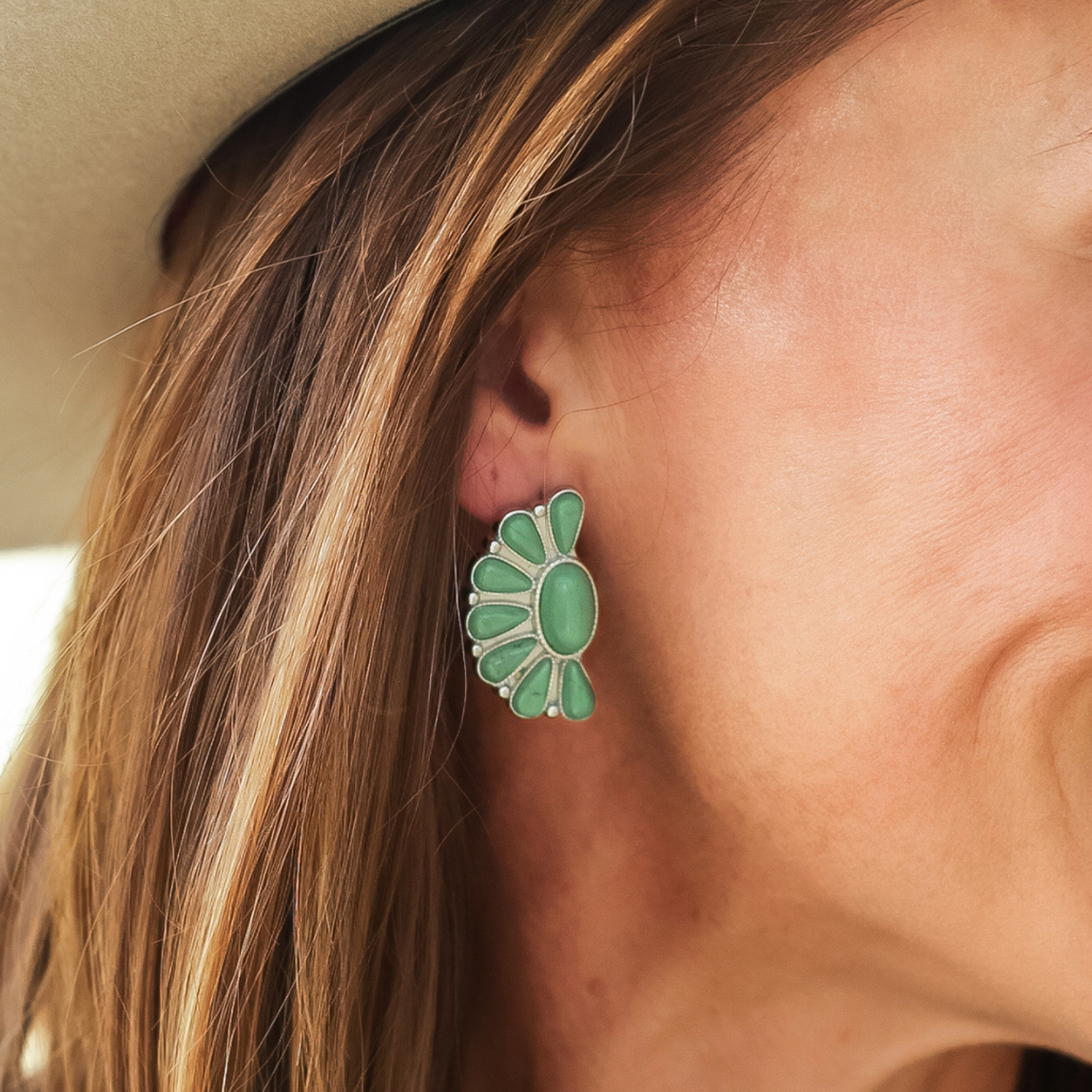 Close-up of a person wearing The Fern Stud Earrings, which are large, green, and fan-shaped with multiple gemstones arranged in a semicircular pattern reminiscent of Western jewelry. Part of their hair and a beige hat are visible.