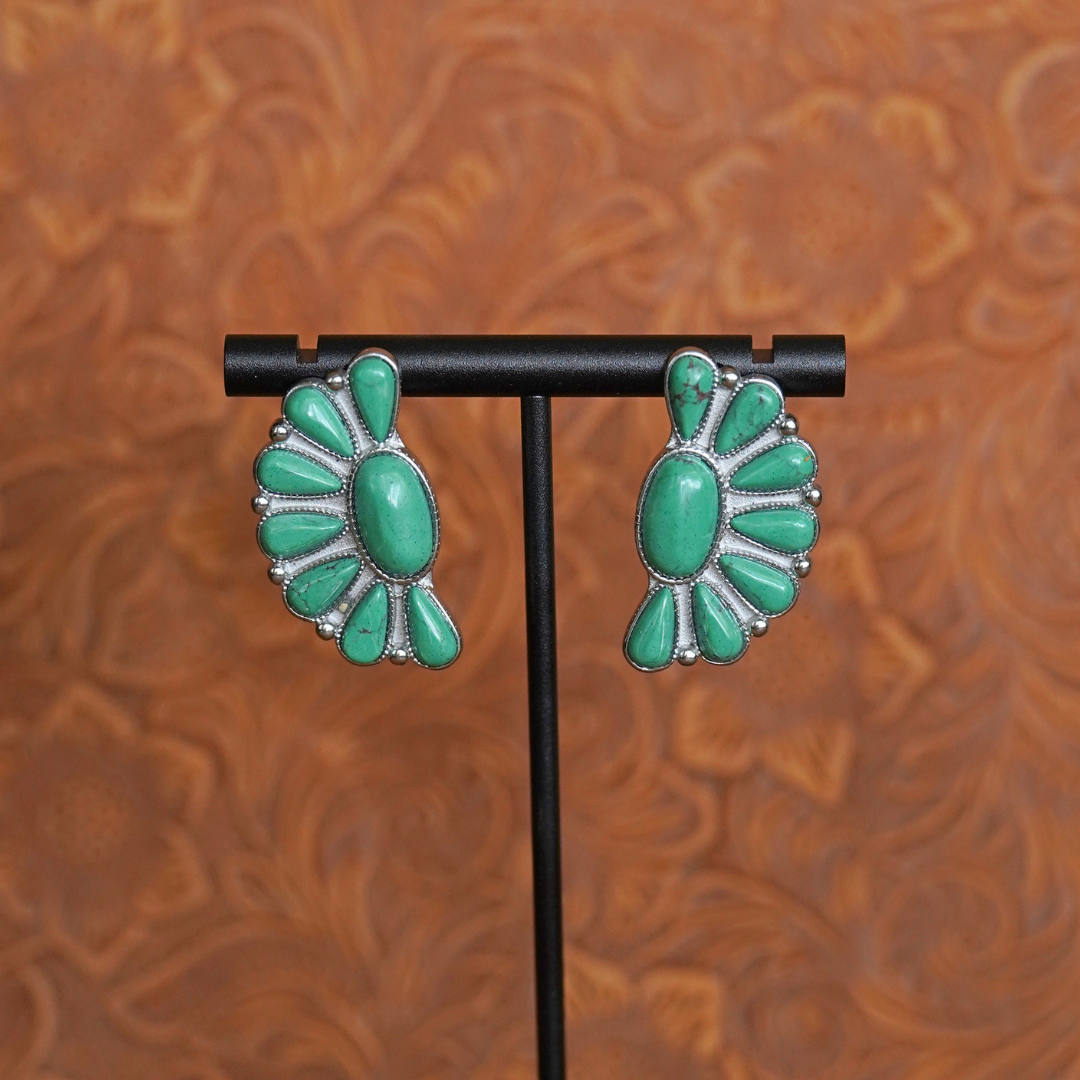 A pair of “The Fern Stud Earrings” in green turquoise with a fan-like design sits elegantly on a black stand, set against a brown, patterned background—a striking example of Western jewelry.
