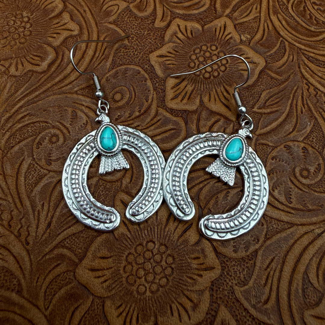 A pair of The Glenna Drop Earrings—ornate silver statement pieces featuring crescent-shaped designs and vibrant turquoise stones—displayed on a textured brown leather surface embossed with floral patterns.