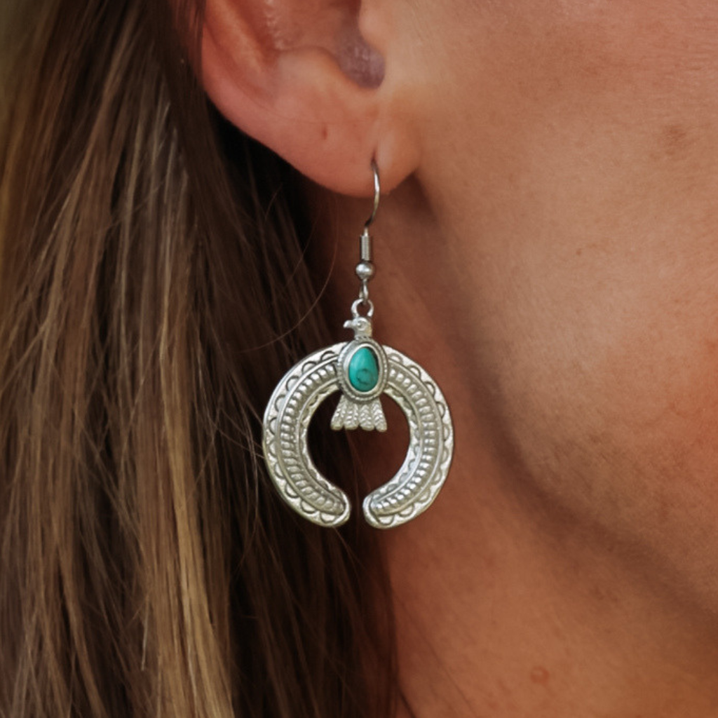 A close-up of a person wearing The Glenna Drop Earrings—silver crescent-shaped earrings adorned with a turquoise stone—against their ear and brown hair; these striking turquoise earrings make the perfect statement accessory for any look.