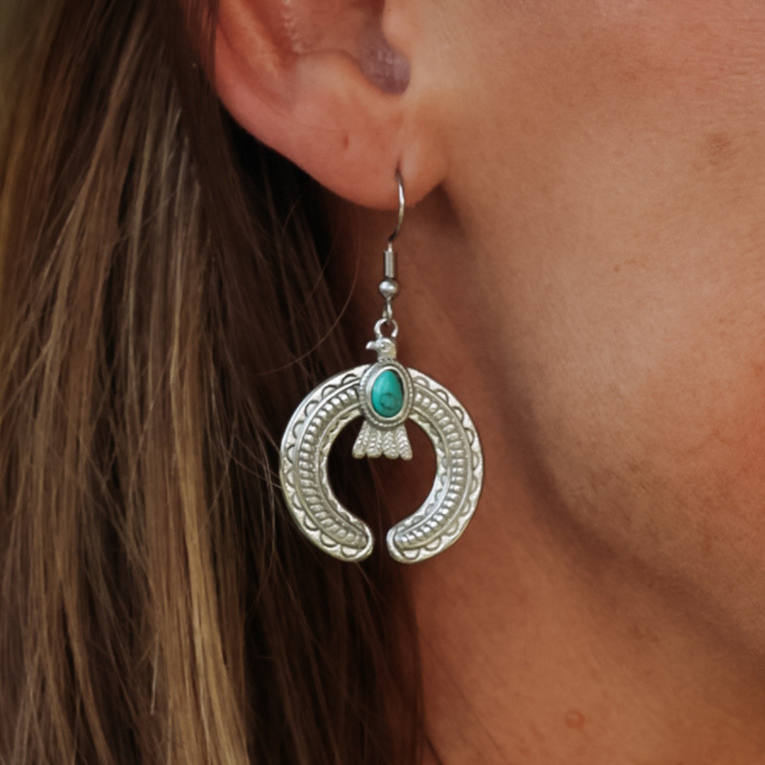 A close-up of a person wearing The Glenna Drop Earrings—silver crescent-shaped earrings adorned with a turquoise stone—against their ear and brown hair; these striking turquoise earrings make the perfect statement accessory for any look.