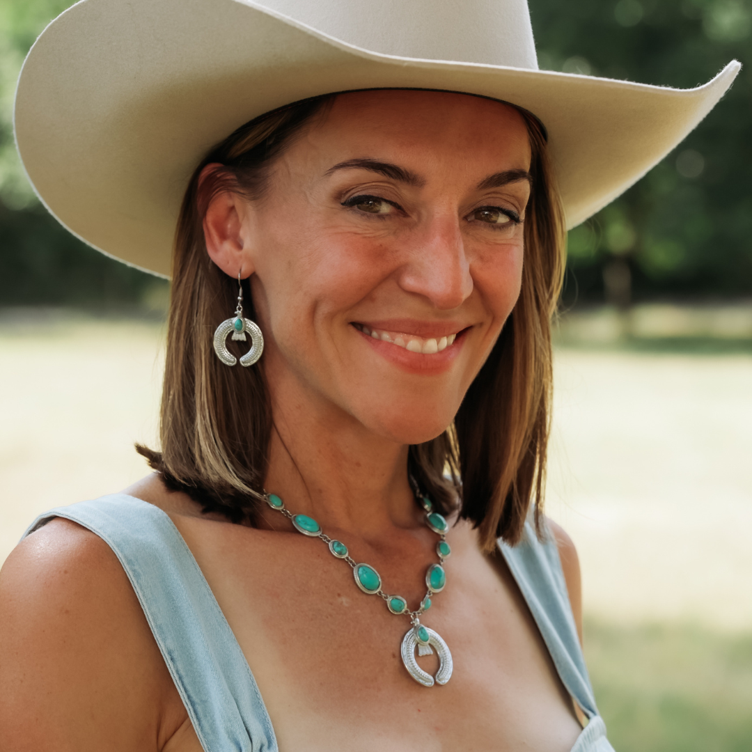 A smiling woman in a wide-brimmed hat, turquoise necklace, and The Glenna Drop Earrings stands outdoors on a sunny day with green trees in the background.