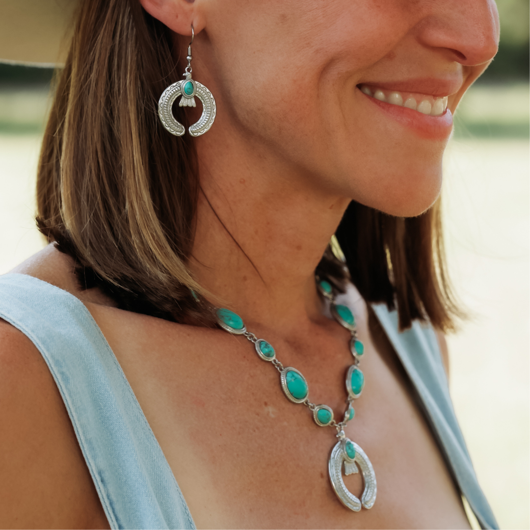 A woman smiles while wearing The Glenna Drop Earrings—striking turquoise earrings that complement her silver and turquoise statement necklace. She has shoulder-length brown hair and is dressed in a light blue sleeveless top.