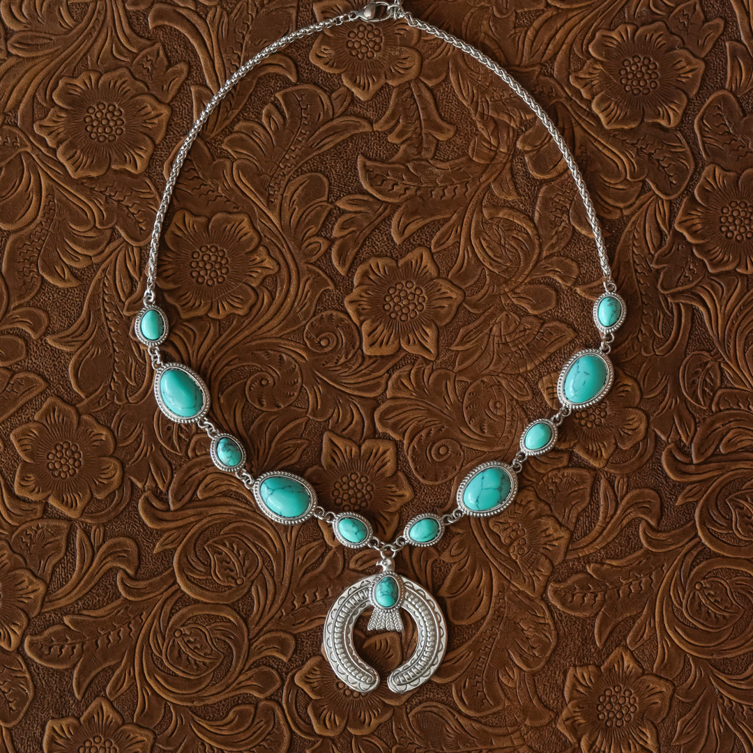 A silver necklace called The Glenna with turquoise stones is displayed on embossed brown leather with a floral pattern.