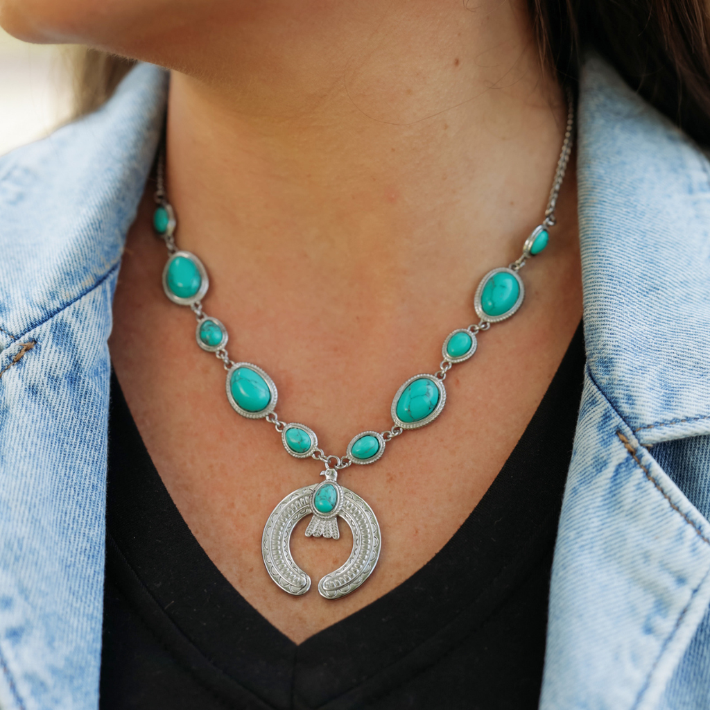 A person wearing The Glenna—a striking turquoise and silver necklace featuring a crescent-shaped pendant—dressed in a black top and light blue denim jacket.