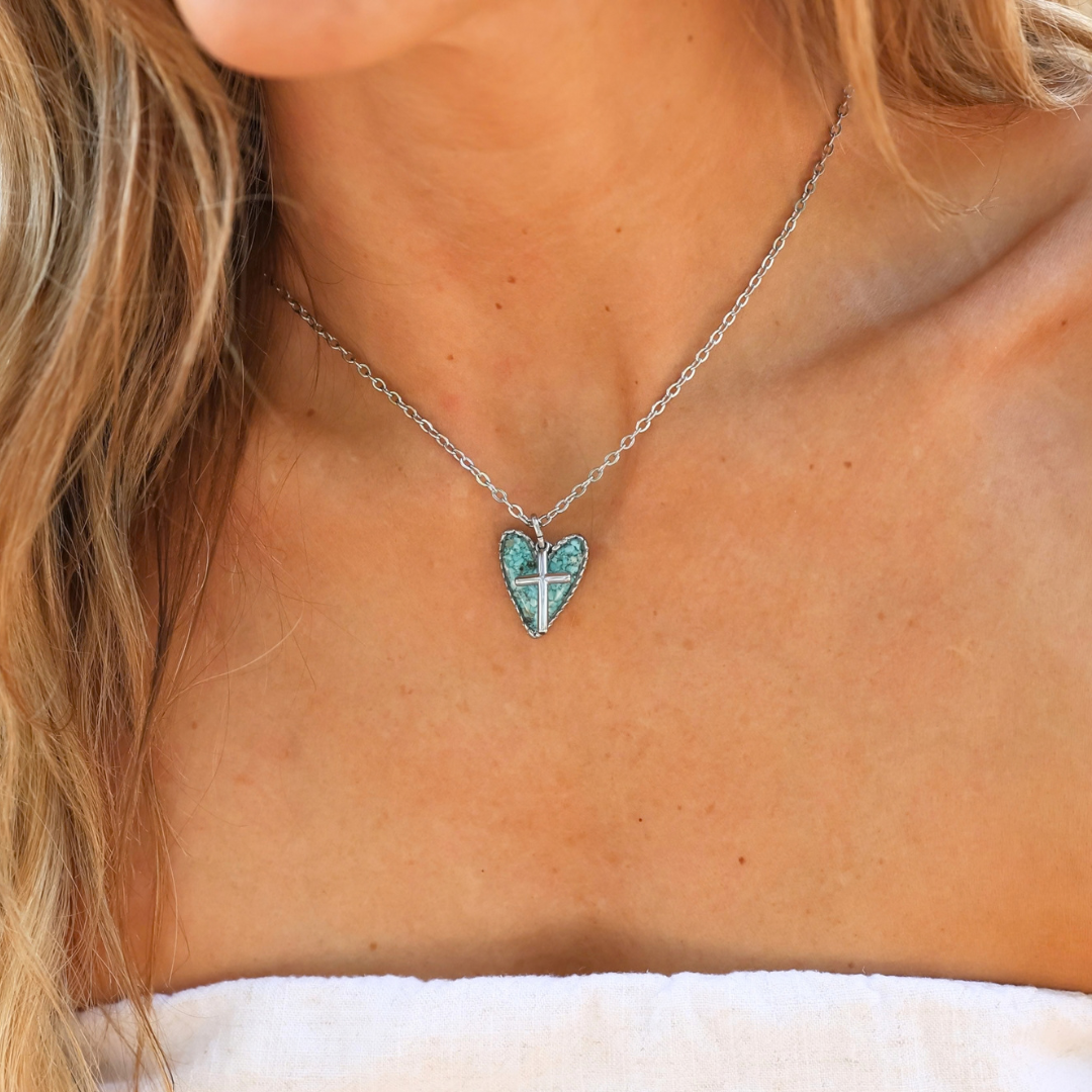A person wearing The Grace, a silver chain featuring a heart-shaped turquoise pendant and a silver cross.