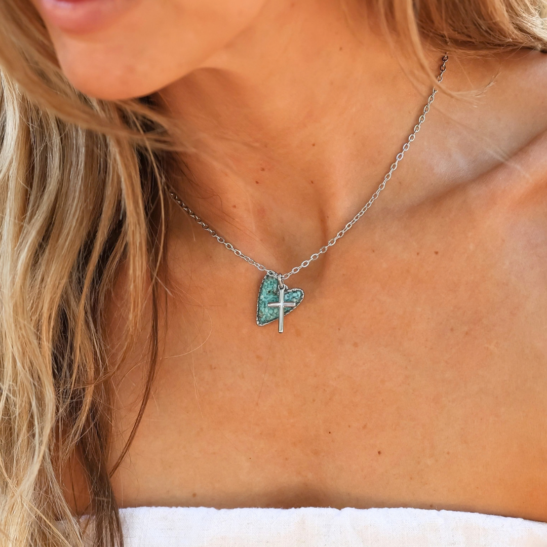 The Grace is a silver chain necklace featuring a small, textured, turquoise heart pendant with a silver cross layered on top, perfect for Western jewelry styles.