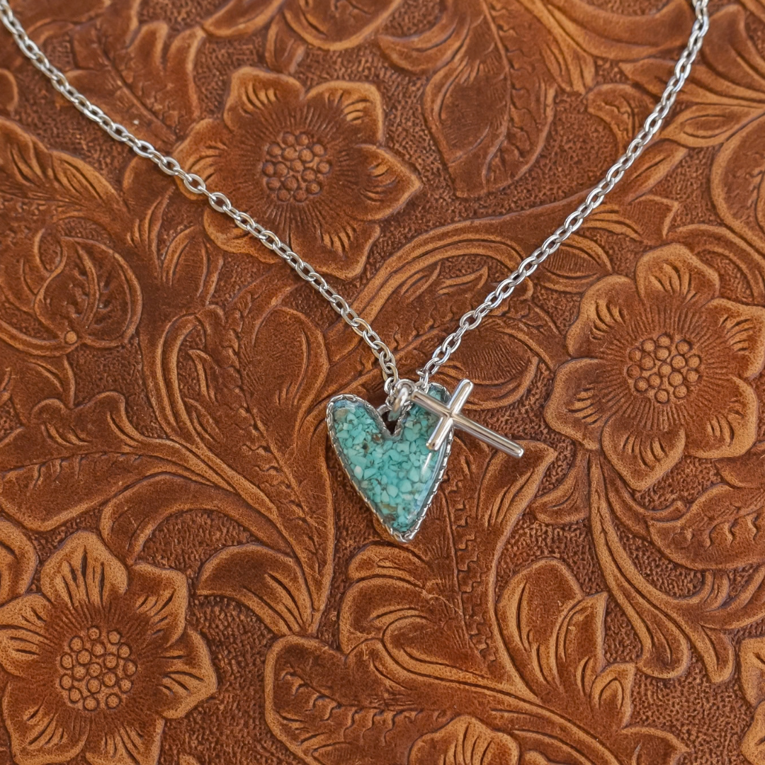 The Grace necklace, a silver chain featuring a heart-shaped turquoise pendant and a small silver cross charm.