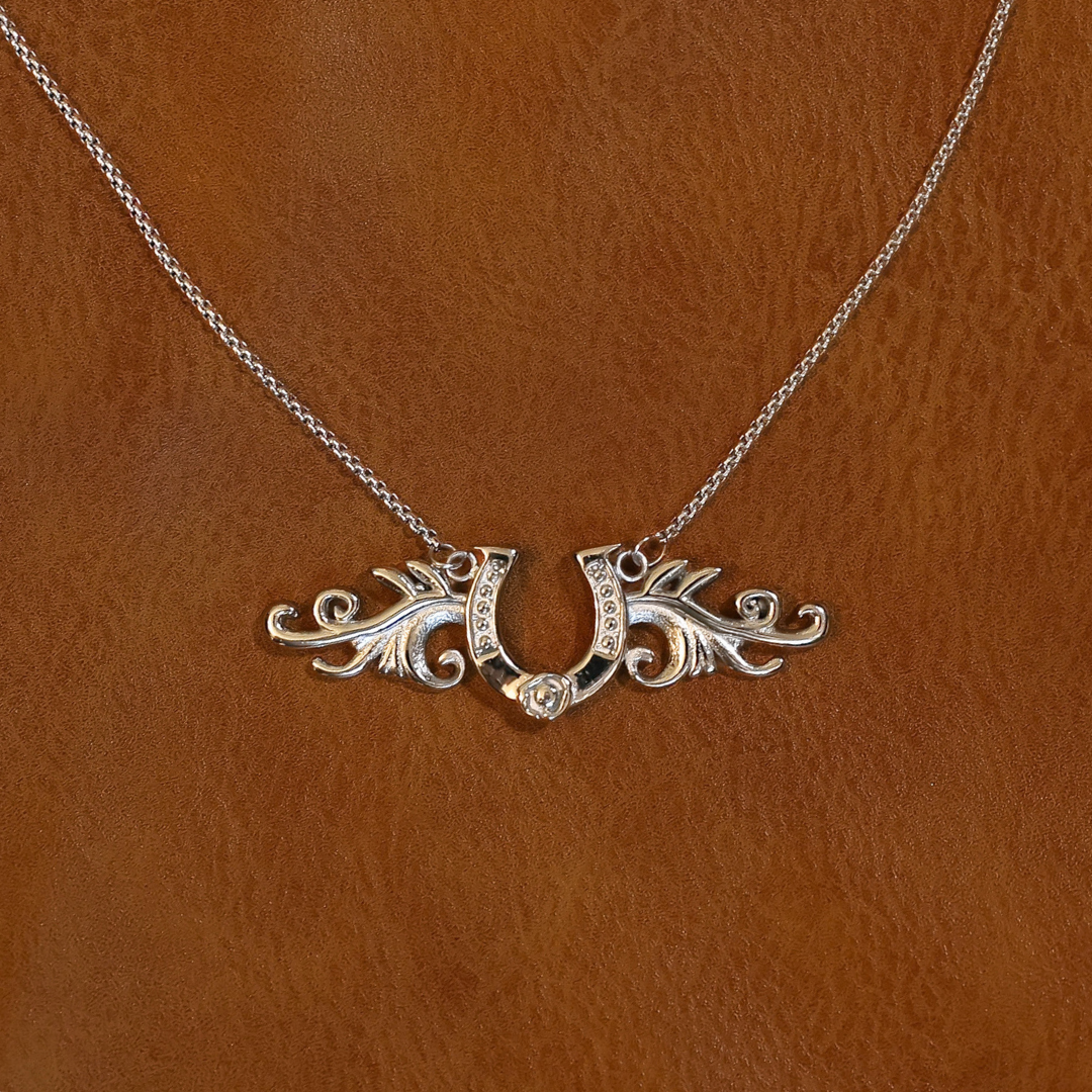 The Gretel is a silver horseshoe pendant necklace featuring filigree wing accents and embedded stones, a classic lucky design.