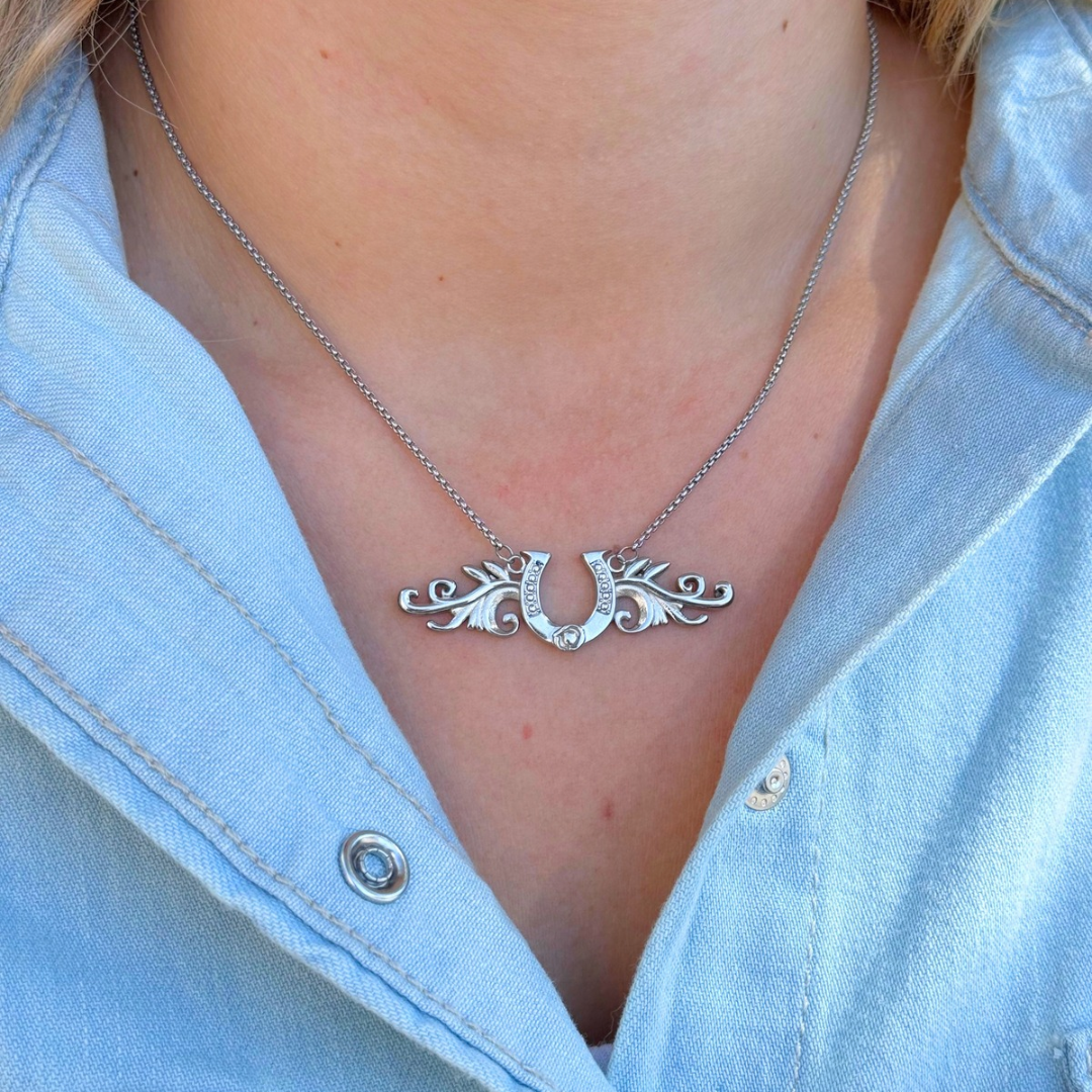 The Gretel, a silver horseshoe necklace with ornate wings on a delicate chain, is worn around a person’s neck as a lucky pendant.