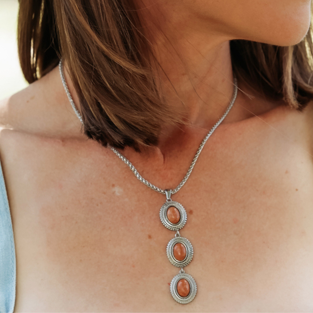 A close-up of a woman wearing The Hallie, a silver Western necklace featuring three oval orange gemstones each set in an ornate silver frame. Her brown hair is shoulder-length, and she’s dressed in a light-colored top.