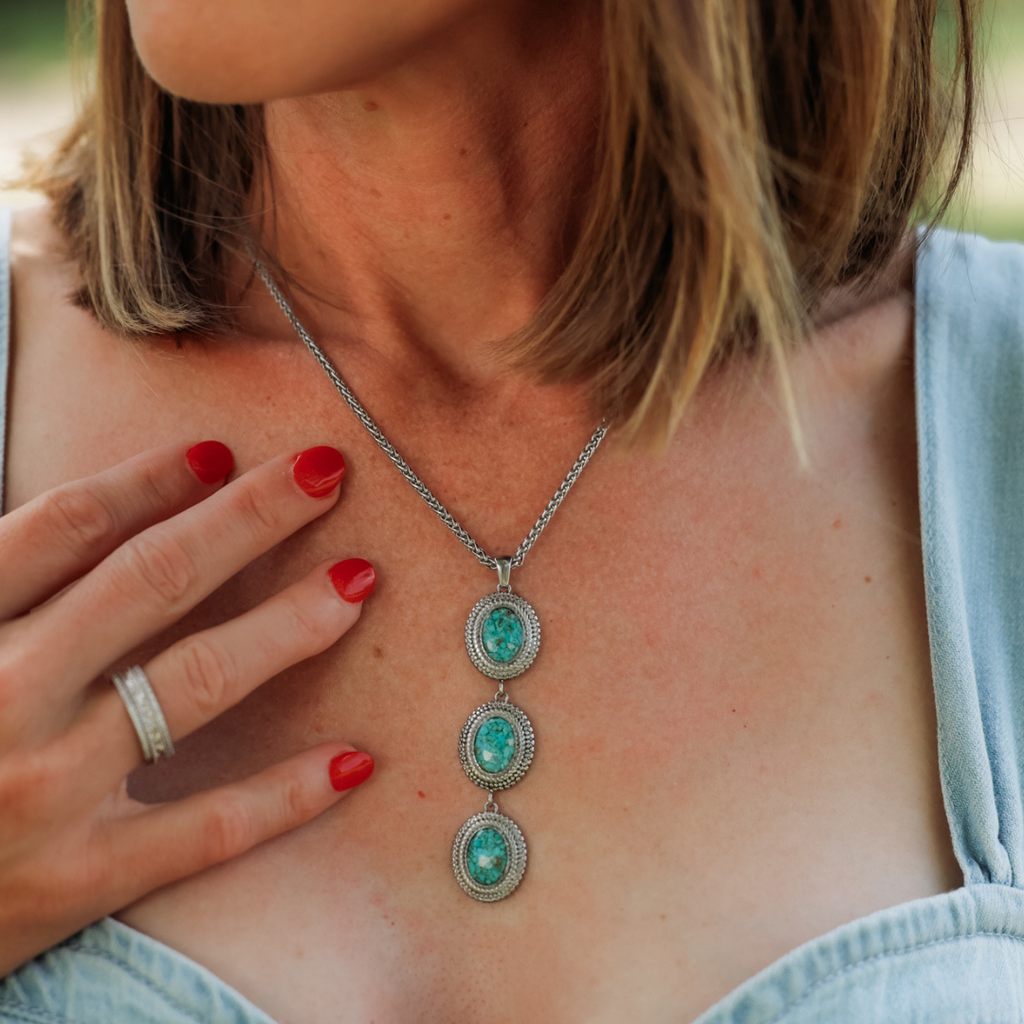 A woman with short brown hair wears The Hallie, a silver necklace accented with three turquoise pendants. Her hand, featuring red-painted nails and a silver ring, rests on her chest as she poses in a light blue top.