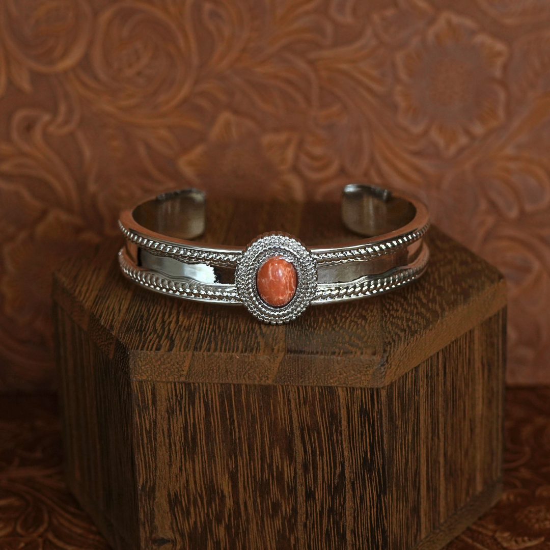 The Hallie Cuff—a silver Western bracelet featuring braided edges and an oval orange stone centerpiece—rests on a hexagonal wooden stand against a brown floral-patterned background, making for striking statement jewelry.