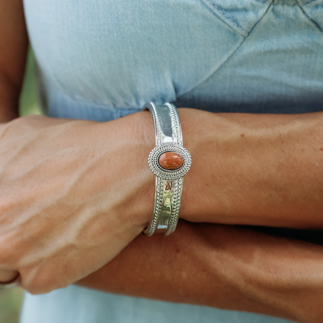 A person wearing The Hallie Cuff—a statement Western bracelet featuring an oval reddish-brown stone at its center—stands with arms crossed, dressed in a light blue top.