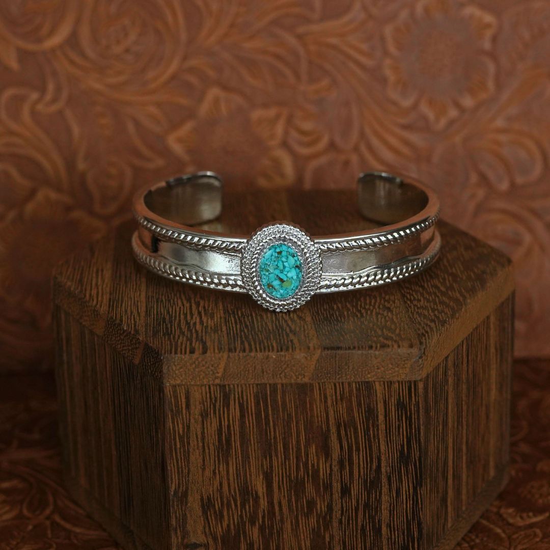 Introducing The Hallie Cuff—a stunning Western-inspired bracelet showcasing a bold turquoise centerpiece set in silver. Elegantly displayed on a wooden box against a brown, floral-patterned backdrop, this eye-catching piece is the perfect statement accessory for jewelry enthusiasts.