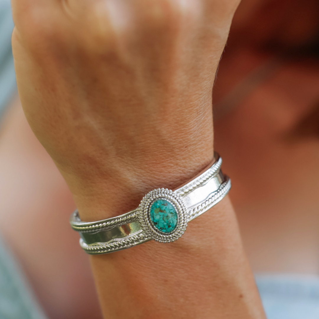 Wearing The Hallie Cuff, a striking bracelet adorned with an oval turquoise stone and intricate detailing around both the stone and edges, makes for a perfect statement jewelry piece suitable for any occasion.