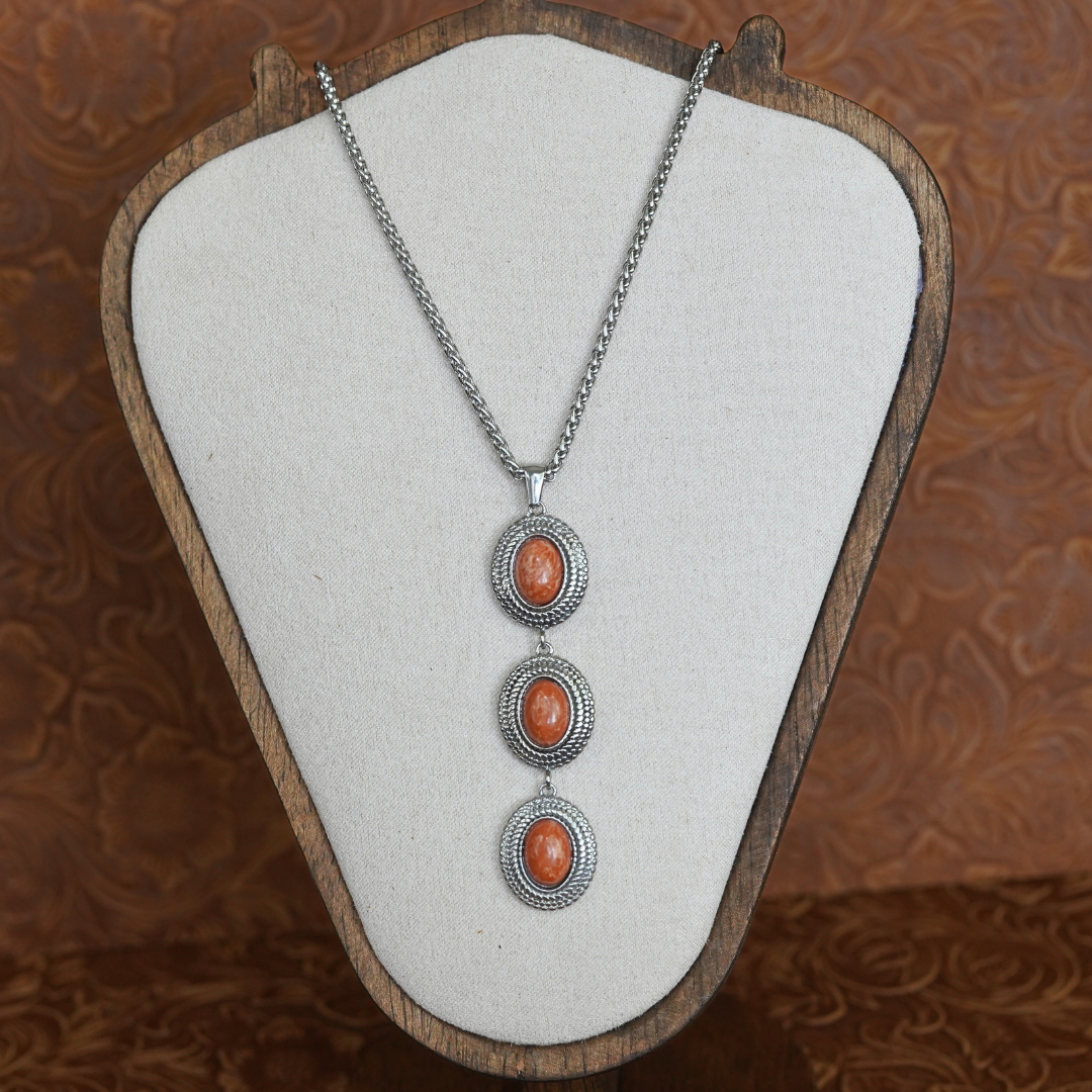 The Hallie necklace features three oval orange stones set in decorative silver frames, inspired by classic Western designs, and is shown displayed on a beige jewelry stand against a brown patterned background.