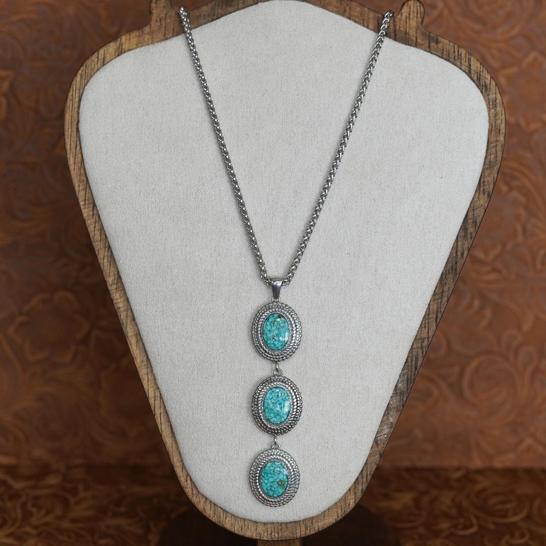 Displayed on a beige jewelry stand with a wooden frame against a brown patterned background, The Hallie showcases three turquoise oval pendants accented by small clear stones on a silver Western-inspired necklace. This striking piece exemplifies timeless style and Southwestern flair.