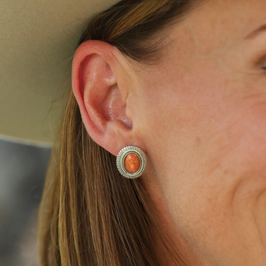 A close-up of a woman’s ear wearing The Hallie Studs—round earrings featuring an orange gemstone, framed by a textured silver border with subtle rope-inspired detailing. She has light skin, light brown hair, and is wearing a light-colored hat.