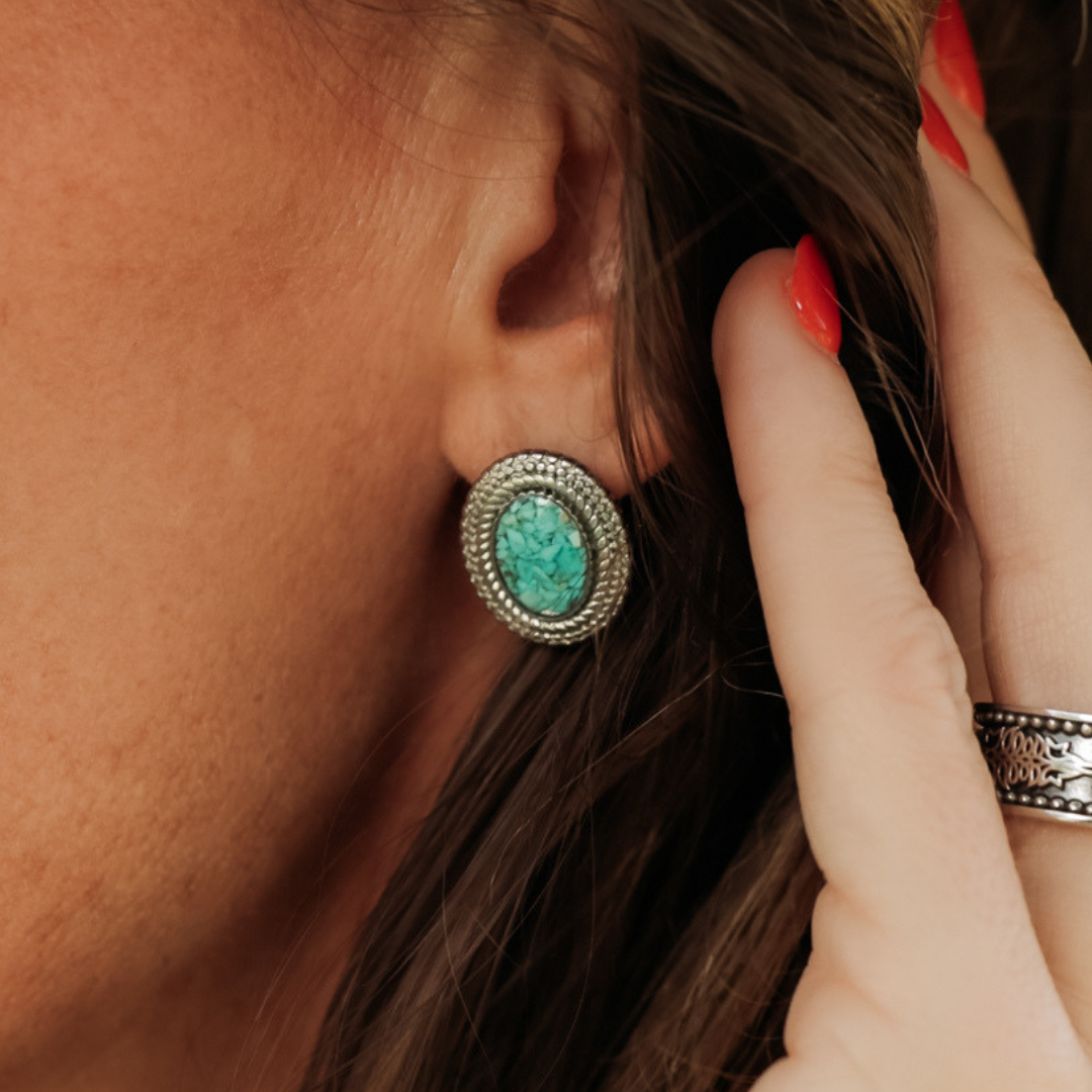 A close-up of a person wearing The Hallie Studs—oval turquoise and silver earrings featuring subtle rope-inspired accents. The person’s hand, adorned with red nail polish and a silver ring, is touching their hair near the ear—an elegant showcase of these distinctive Western studs.