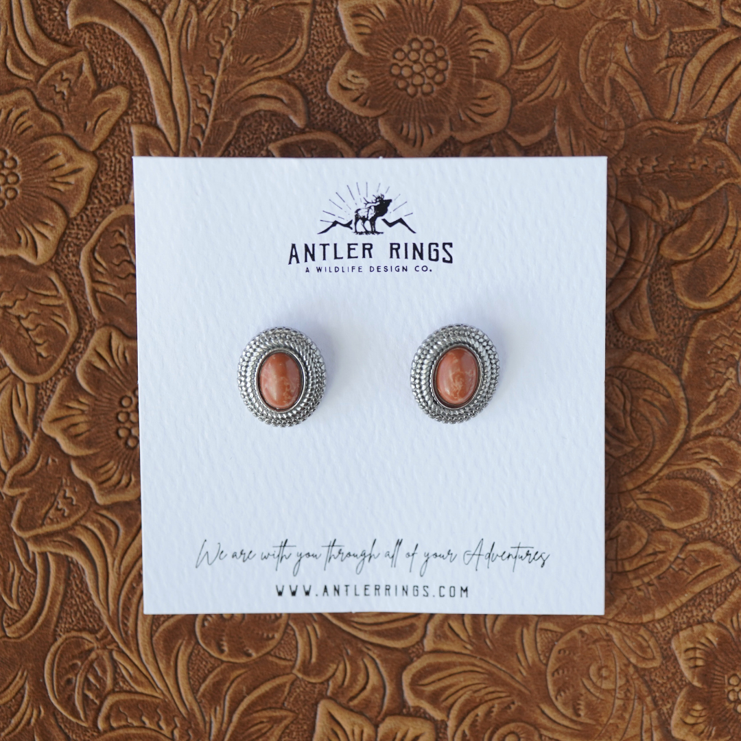 A pair of The Hallie Studs—oval Western earrings featuring brown stone centers and subtle rope-inspired accents—rests on a textured white card labeled “Antler Rings,” displayed atop a brown, floral-patterned surface.
