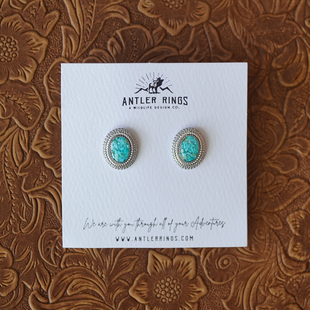 A pair of The Hallie Studs—oval turquoise stud earrings featuring rope-inspired accents and silver detailing—displayed on a branded Antler Rings card against an embossed brown floral surface.