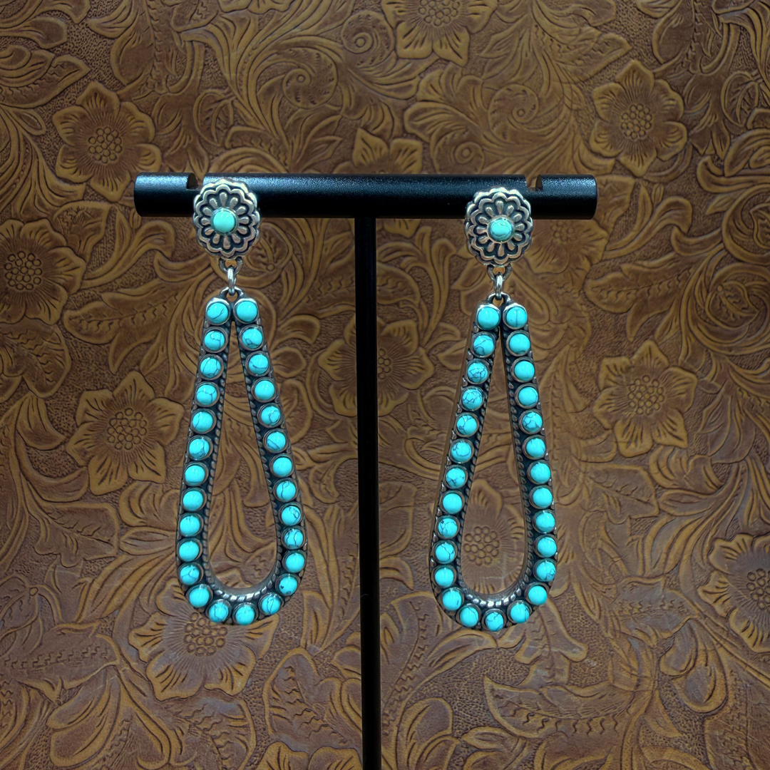 The Heidi Drop Earrings feature teardrop-shaped turquoise stone loops and a floral silver stud, displayed on a stand against a brown floral-patterned background.