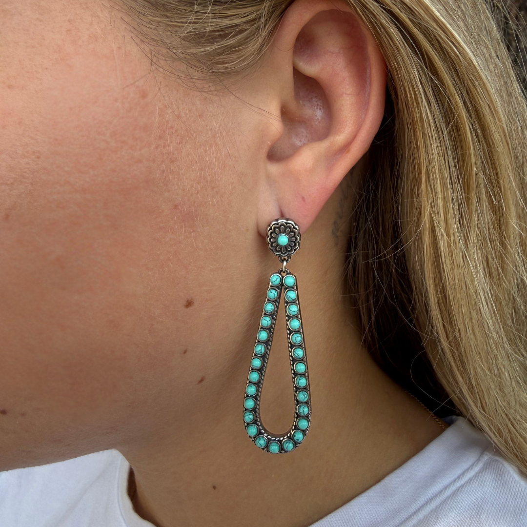 A close-up of a woman’s ear showcases The Heidi Drop Earrings—a striking pair featuring large teardrop-shaped turquoise drops and a silver floral stud crafted from stainless steel. Her light blonde hair frames the earring, creating a modern take on classic Western style.