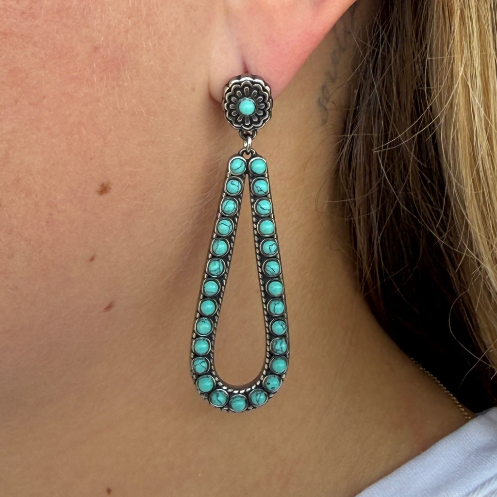 A close-up of a person’s ear wearing The Heidi Drop Earrings—a teardrop-shaped design featuring turquoise stones set in silver. The earring has a floral stud and a loop adorned with vibrant turquoise gems, capturing the essence of classic turquoise drop earrings. Part of the person’s blond hair is visible.
