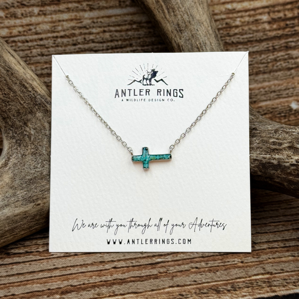 Introducing The Hope: a turquoise cross necklace on a silver chain, beautifully displayed on an Antler Rings card with antlers in the background. The card reads, “We are with you through all of your Adventures.” This stunning necklace adds a unique touch to any outfit while symbolizing strength and faith.