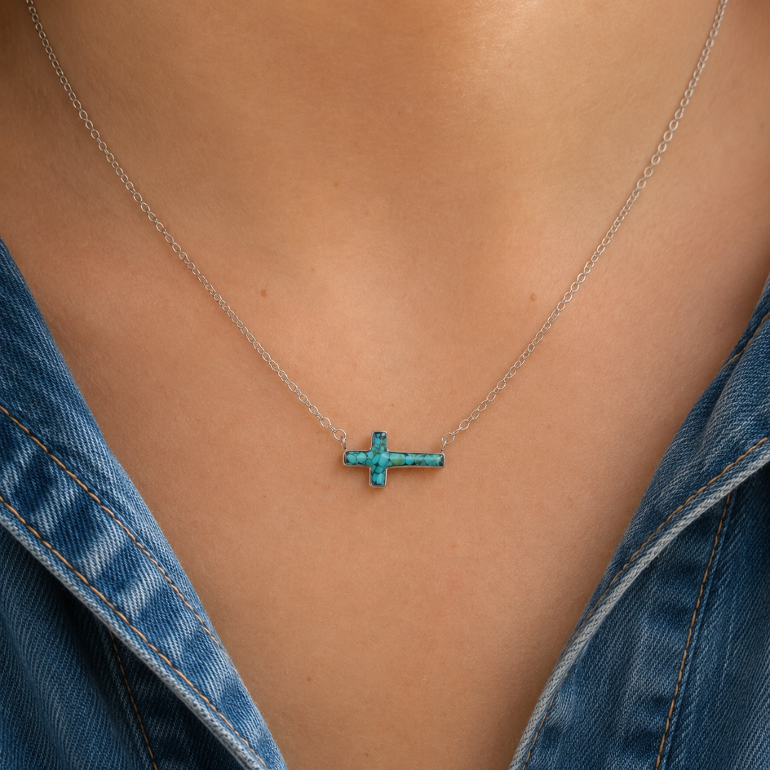 A close-up of a person wearing The Hope, a stainless steel cross necklace with a horizontal turquoise pendant, visible against their skin and a blue denim shirt.