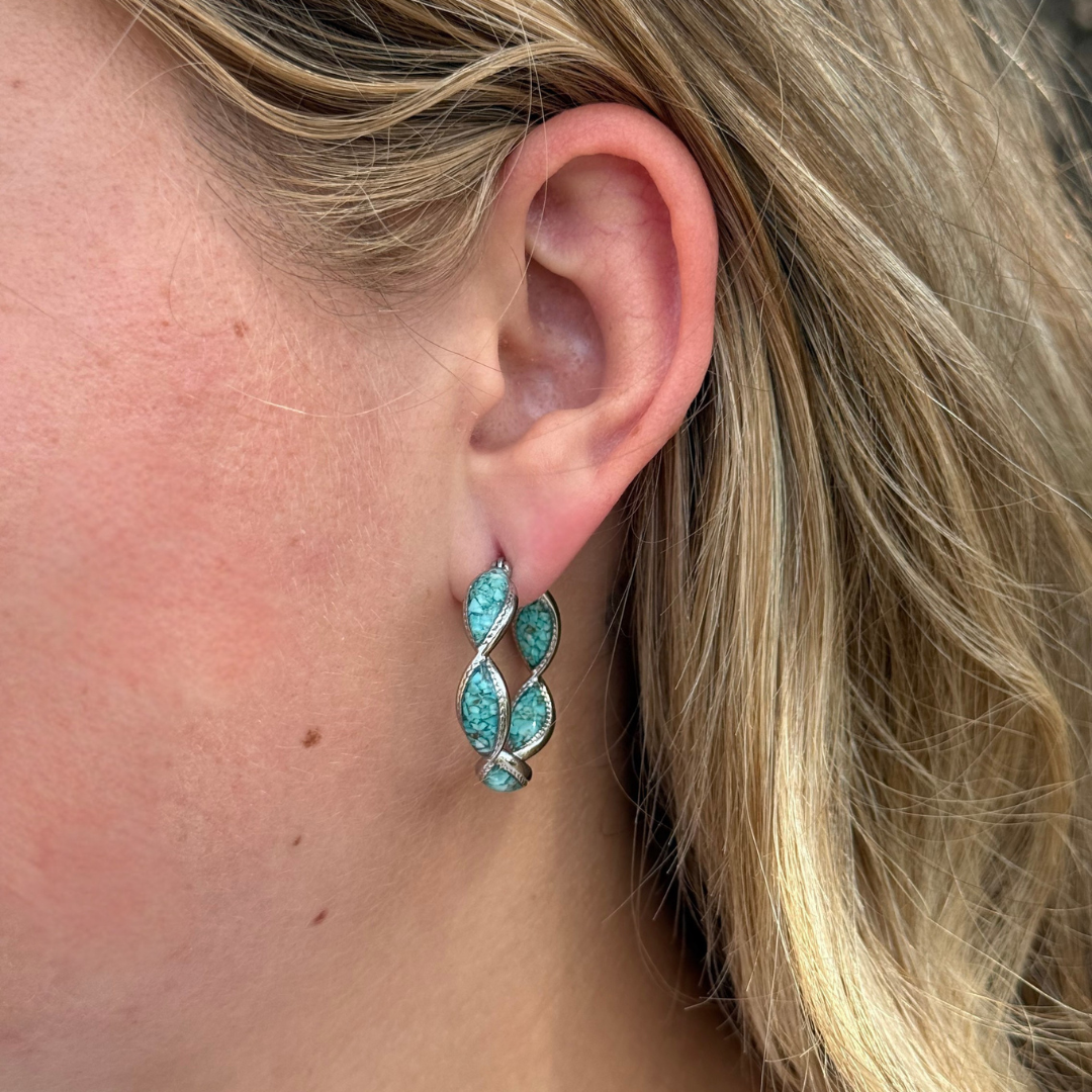 A close-up of a woman’s ear wearing The Janell Hoops—silver earrings featuring four blue-green, leaf-shaped stones in a Western style design. Blonde hair is visible, and the background is out of focus.