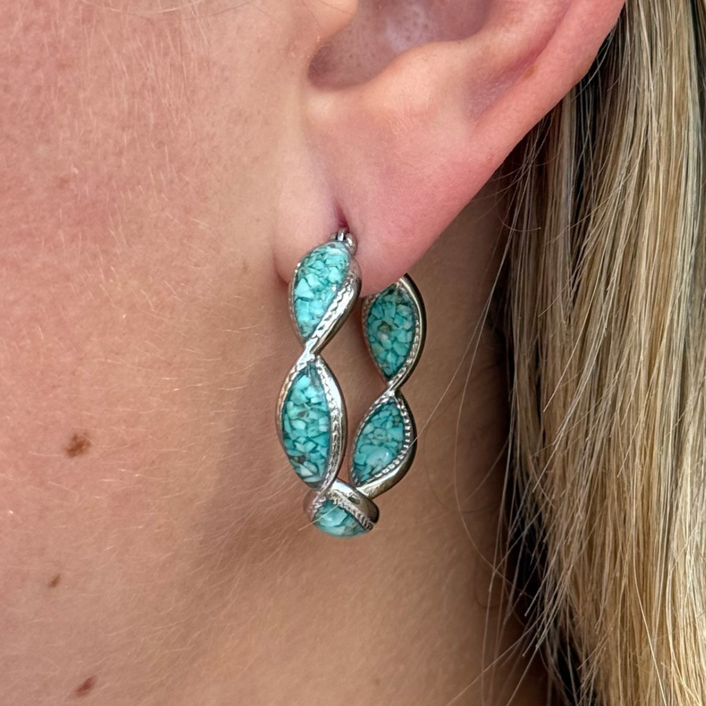 A close-up of an ear wearing The Janell Hoops—silver earrings featuring three crushed turquoise stones set in a twisted, leaf-like design. The wearer has light skin and blonde hair, highlighting the Western style of this jewelry.