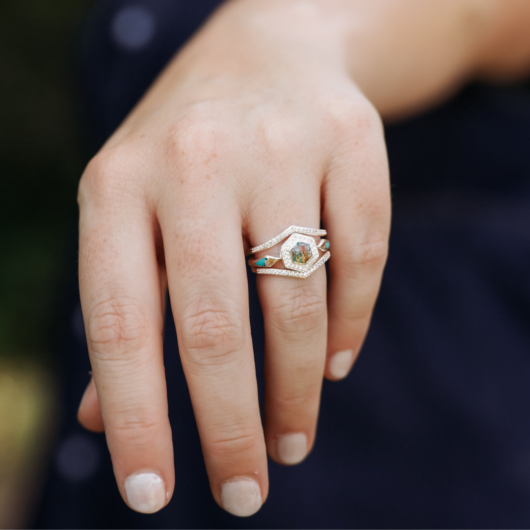 A hand with neatly trimmed nails wears The Jolene, a geometric multi-stone Spiny Oyster turquoise ring featuring gold and turquoise details, set against a blurred dark background.