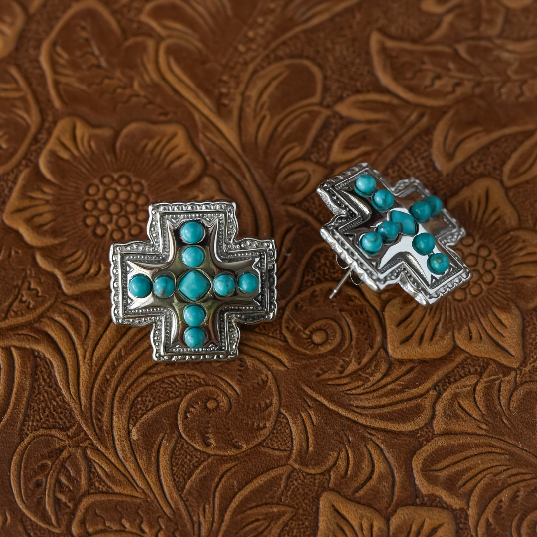 The Kalyn Studs—two silver cross-shaped earrings featuring turquoise stones—rest on brown embossed leather with a floral pattern, perfectly capturing the spirit of Western jewelry.