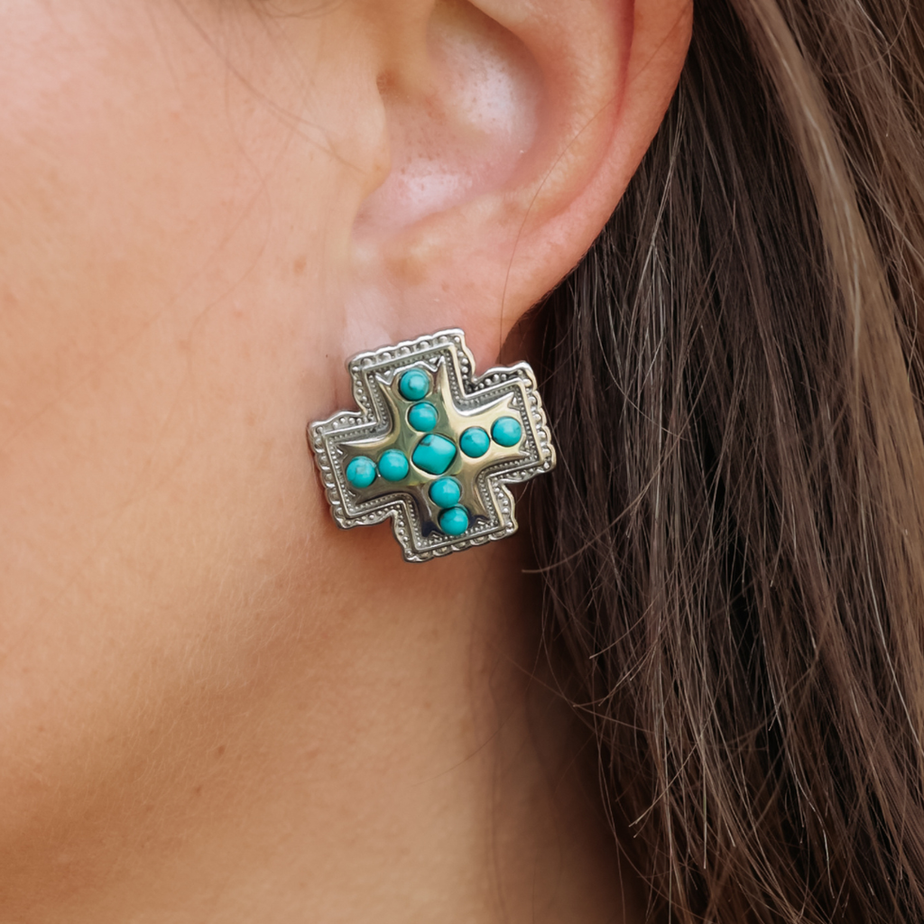 A close-up of a person’s ear wearing The Kalyn Studs—large, silver cross-shaped earrings adorned with turquoise stones arranged in the center and along the arms of the cross. The striking turquoise cross earring stands out as a statement piece of Western jewelry, while brown hair gently frames the ear.