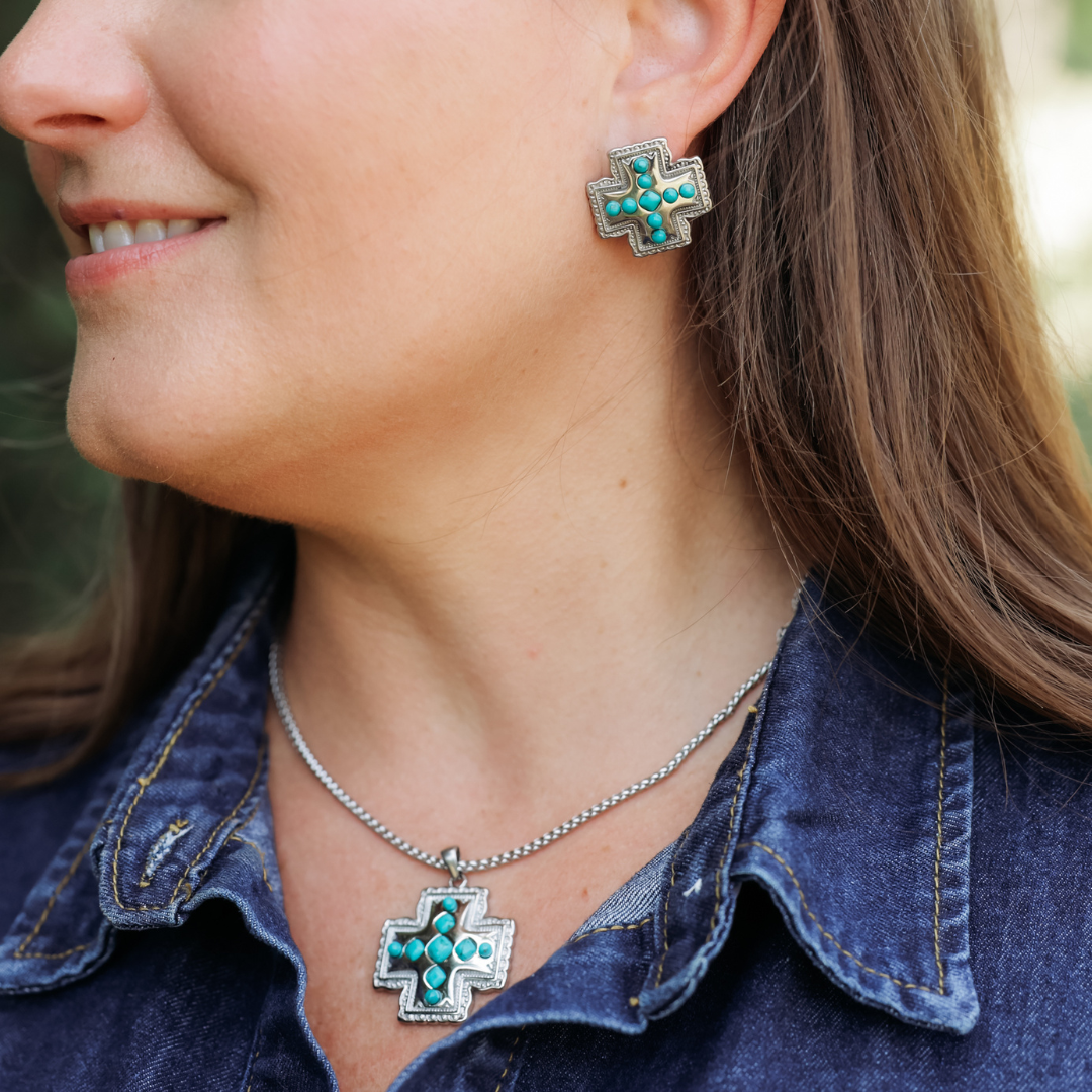 Woman wearing a denim shirt and Western jewelry featuring The Kalyn Studs—silver and turquoise cross-shaped faith earrings—as well as a matching necklace, smiling slightly and facing left.