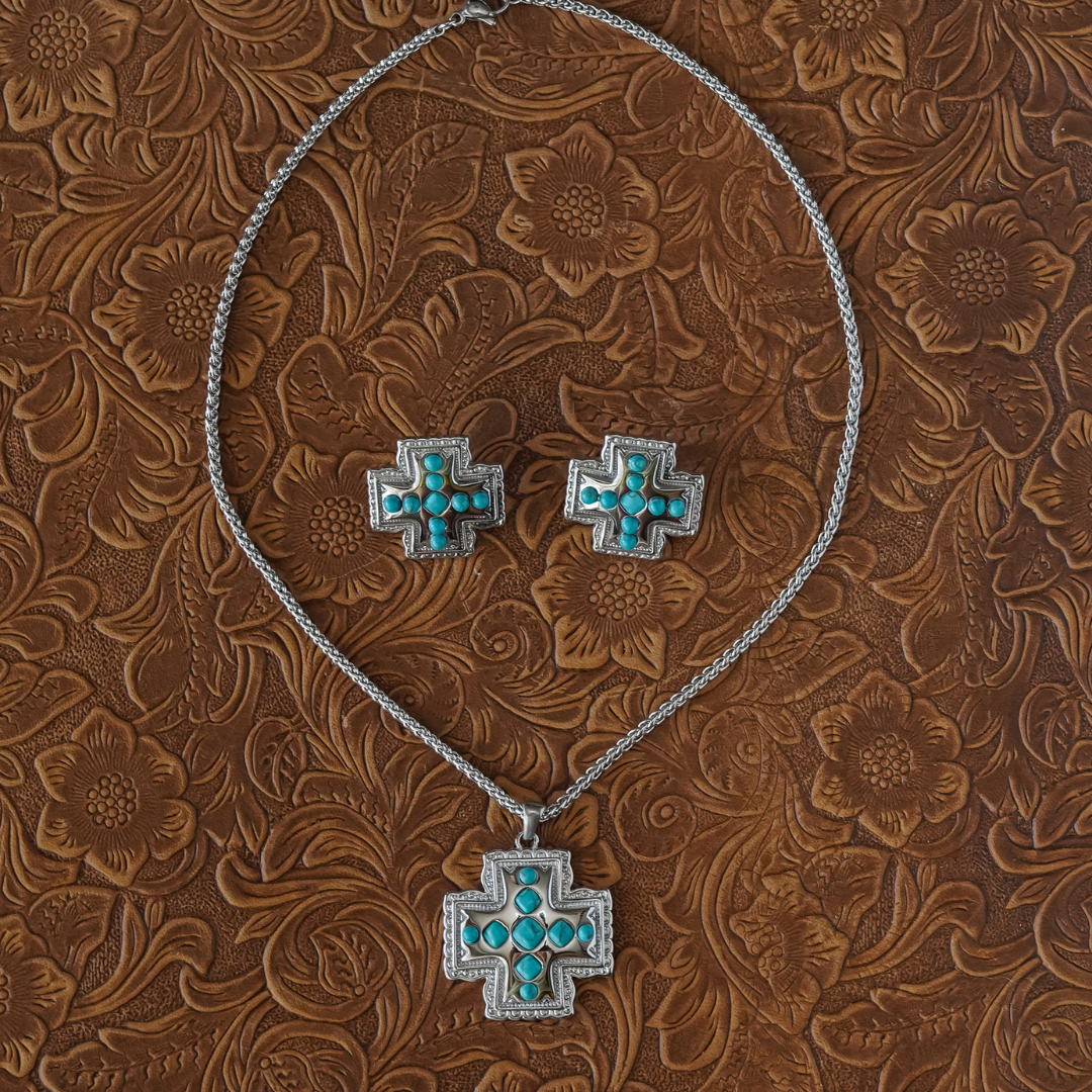 The Kalyn Studs—silver earrings featuring cross-shaped turquoise stone inlays—are displayed on a brown, floral-embossed leather surface, showcasing a striking example of Western jewelry.