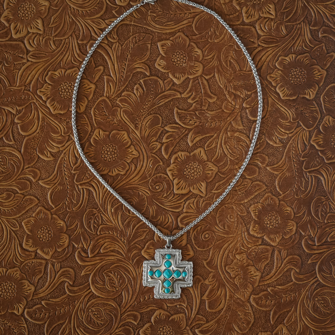 The Kalyn—a striking turquoise cross necklace featuring vibrant turquoise stones set in a silver cross pendant on a sleek silver chain—stands out against a brown, floral tooled leather background, making it an elegant piece of Western jewelry that beautifully celebrates faith and style.